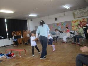フルート奏者の前で手をつないで踊る先生と子ども